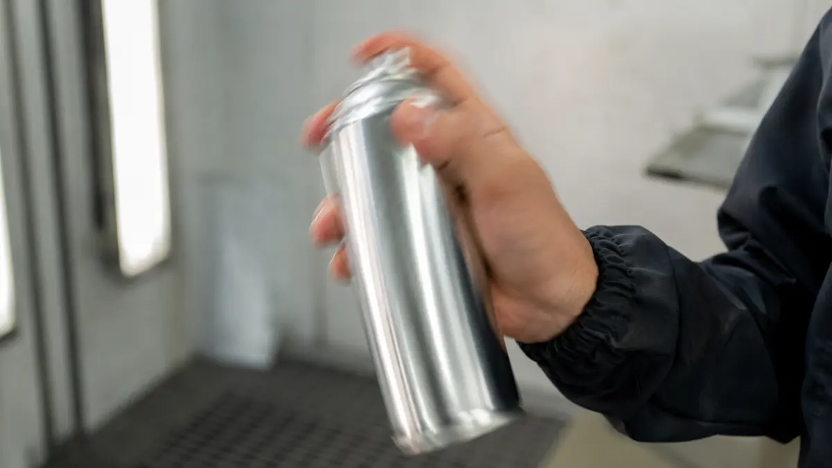 Hand schüttelt eine Chromlack-Sprühdose senkrecht vor Werkstatthintergrund